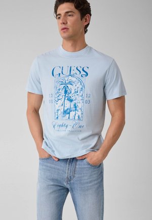 Giovane uomo che indossa una T-shirt Guess azzurra con grafica di una palma e jeans blu, in piedi con le mani nelle tasche davanti a uno sfondo semplice.