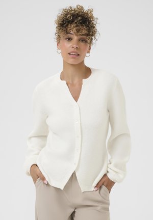 Weißer gestrickter Cardigan mit rundem Ausschnitt, Knopfleiste, langen Ärmeln und einer weichen Textur. Tragen mit beigen Hosen.