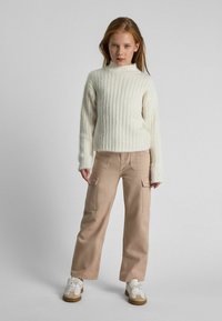 Cremefarbener gerippter Strickpullover mit hohem Kragen, kombiniert mit beigen Cargo-Hosen mit Seitentaschen. Schuhwerk: weiße Sneakers mit braunen Akzenten.