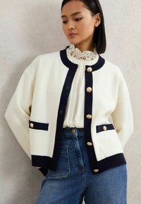 Bela in mornarsko modra kırtka z zlatimi gumbi, rebrastimi manšetami in žepi. Ujemajoča bela bluzica z čipkastim ovratnikom in kavbojke z visokim pasom.