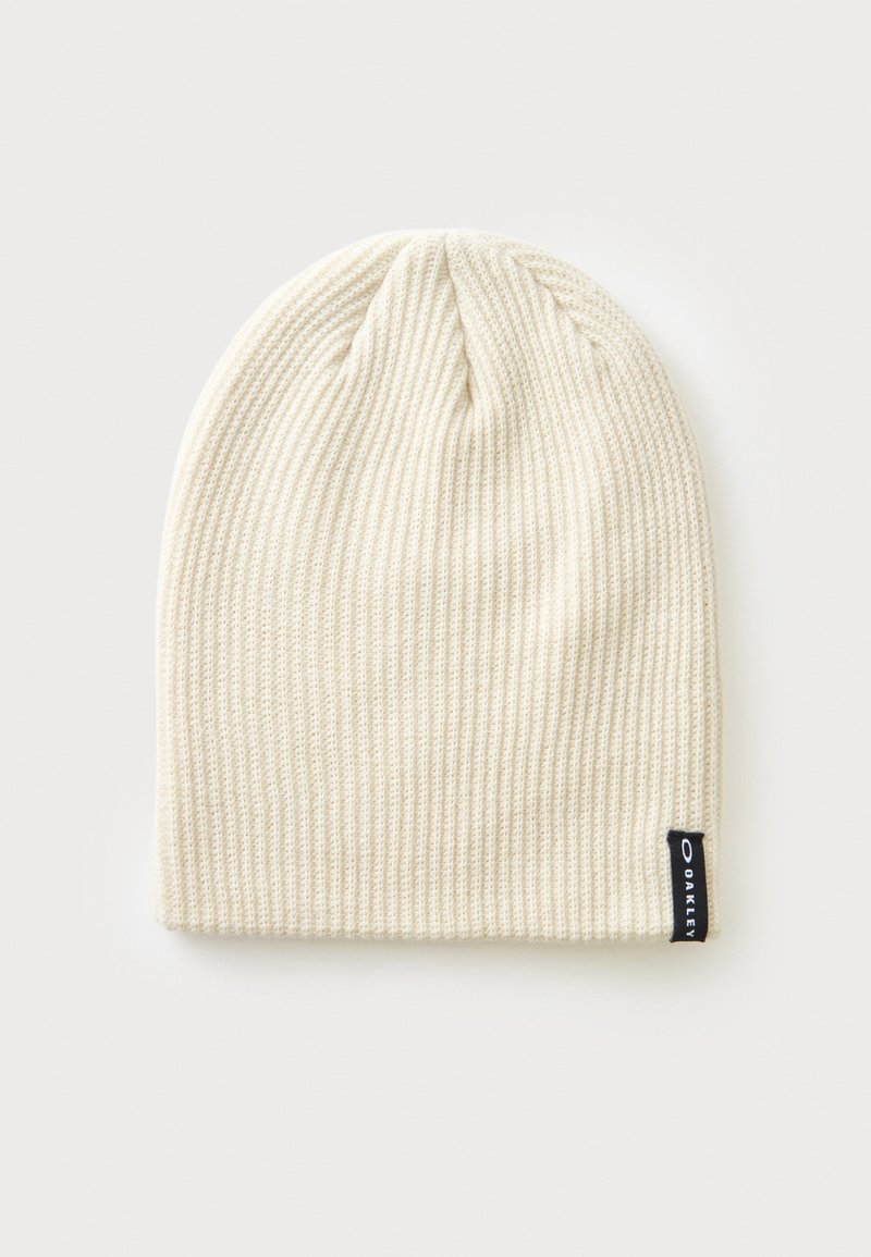 Bonnet en maille côtelée couleur crème avec une petite étiquette noire du logo Oakley sur le bord inférieur, sur un fond clair uni.