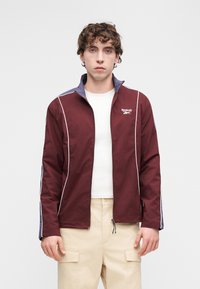 Bordowa kurtka na zamek z białymi akcentami i fioletowym kołnierzem. Wykonana z lekkiego materiału, z logo Reebok na piersi. Nosi się ją na białą koszulę.