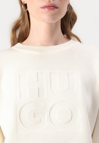 Krémový svetr s žebrovanou texturou a vyraženým logem "HUGO" na přední straně. Obsahuje kulatý výstřih a měkkou tkaninu.