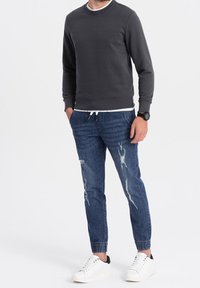 Sudadera gris con cuello redondo, combinada con jeans azules desgastados que tienen puños elásticos. Se lleva con zapatillas blancas y un reloj negro.