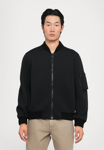 Veste bomber noire avec fermeture éclair à l'avant, col et poignets côtelés, et poche zippée sur la manche gauche. Texture de tissu lisse.