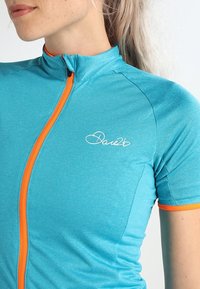 Una camiseta de ciclismo de manga corta en tela turquesa brillante con una cremallera y puños naranjas. Presenta un logo blanco en el pecho.