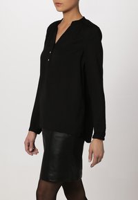 Czarna bluzka z długim rękawem, dekoltem w serek, marszczonymi ramionami i dwoma guzikami przy szyi. Gładki materiał, zestawiona ze skórzaną spódnicą.