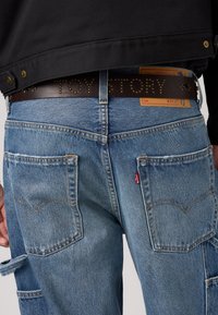 Sinised denim teksad, millel on veidi pleekinud viimistlus, kahe tagataskuga, tugevdatud õmblustega ja pruuni nahkrihmaga, millel on narmastega kirjutatud "TOY STORY."