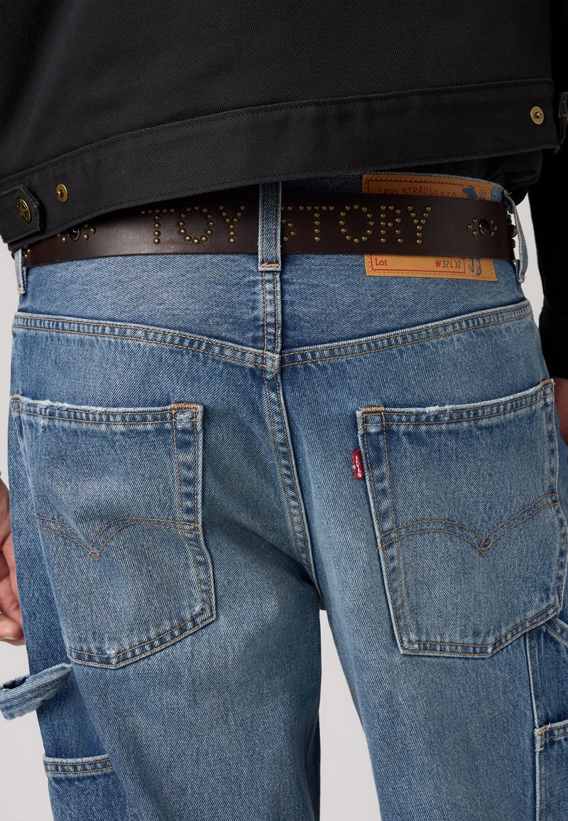 Sinised denim teksad, millel on veidi pleekinud viimistlus, kahe tagataskuga, tugevdatud õmblustega ja pruuni nahkrihmaga, millel on narmastega kirjutatud "TOY STORY."