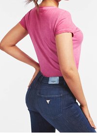 Lyserød bomulds-T-shirt med korte ærmer, kombineret med mørkeblå denimjeans. Jeansene har et trekantet logo og en blå mærkeetiket på bagsiden.