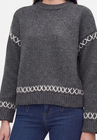 Maglione a maglia grigio scuro con motivi incrociati bianchi su maniche e orlo, indossato con jeans blu, mostrato dal collo ai fianchi.
