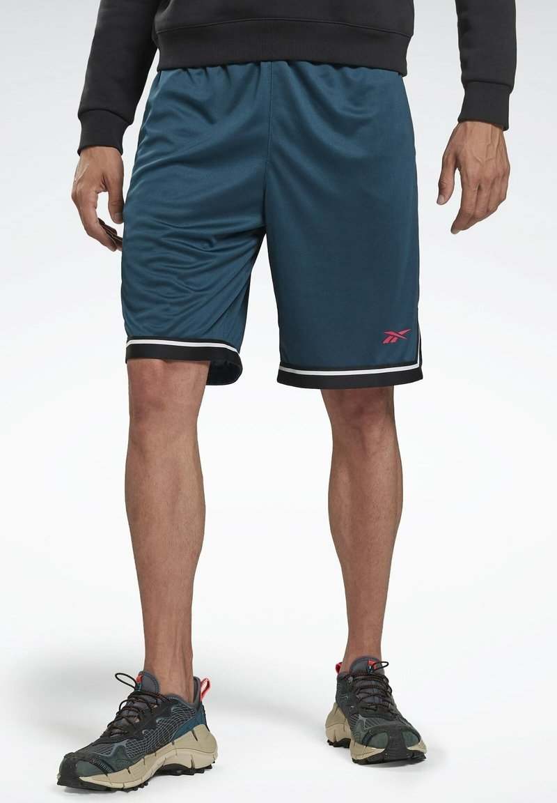 Mann trägt türkisfarbene Sportshorts mit schwarz-weißem Rand, schwarzen Pullover und dunkelgraue Sneakers, steht vor weißem Hintergrund.