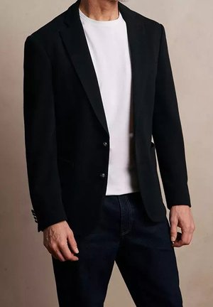 Mann trägt ein schwarzes Sakko über einem weißen T-Shirt und dunklen Jeans, steht vor einem schlichten beigen Hintergrund.