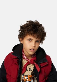 Kind gekleed in een rode puffer vest over een zwarte hoodie, met een crèmekleurig t-shirt met een tijgerafbeelding. Een rode bandana is om de nek geknoopt.