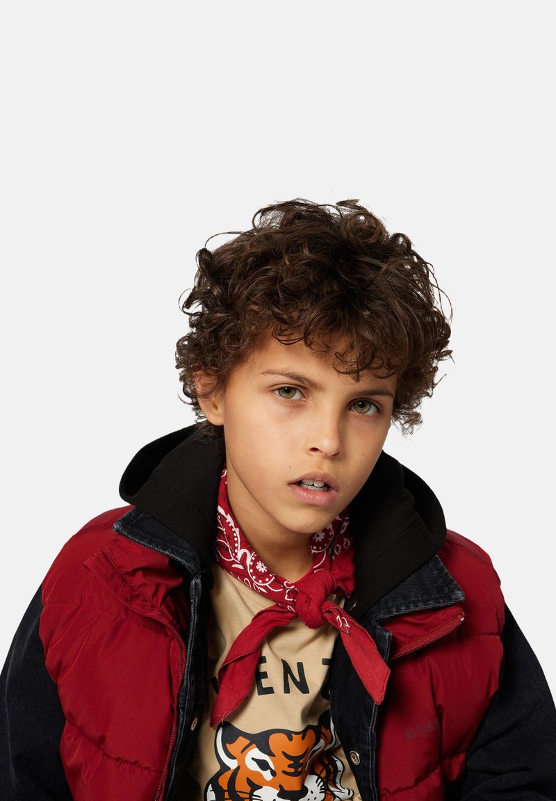 Kind gekleed in een rode puffer vest over een zwarte hoodie, met een crèmekleurig t-shirt met een tijgerafbeelding. Een rode bandana is om de nek geknoopt.