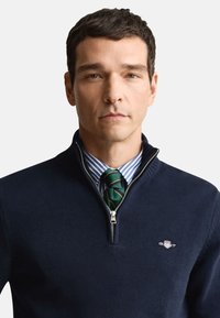 Mornarsko modra pulover z zadrgo, z rebrastim ovratnikom, pleteno teksturo in detajlom logotipa. Nošena preko modre srajce s črtami in zelene kravate s črtami.