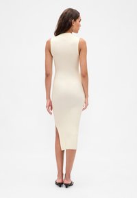 Bordázott, ujjatlan világos bézs ruha oldalsó hasítékkal, testhez simuló sziluettel és magas nyakkal. Fekete magassarkú szandállal viselve.
