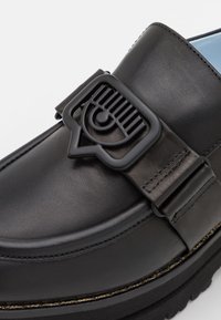 Mocassin en cuir noir avec semelle épaisse, couture détaillée et une boucle noire décorative présentant un motif d'œil stylisé.