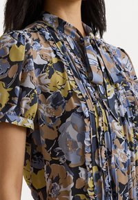 Blusa floreale con maniche corte, dotata di una placca e dettagli arricciati. I colori includono blu, giallo, marrone e nero, con un tessuto leggero.
