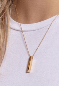 Collana tono oro con un ciondolo rettangolare testurizzato, sospeso da una catena sottile, indossata su una maglietta di colore chiaro.