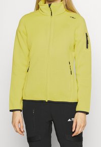 Gelbe Fleecejacke mit hohem Kragen, Frontreißverschluss und zwei seitlichen Reißverschlusstaschen. Mit schwarzen Akzenten und strukturiertem Stoff.