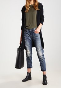 Femme portant un cardigan noir, une chemise vert olive, un jean déchiré bleu, des bottines noires, tenant un grand sac à main noir.