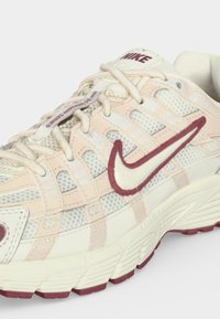 Λευκό και ανοιχτό ροζ αθλητικό παπούτσι Nike, με κόκκινες λεπτομέρειες, δίχτυ σε πάνελ, μαλακή σόλα και το κείμενο "BUCEANAN SERIES" στην ετικέτα των κορδονιών.