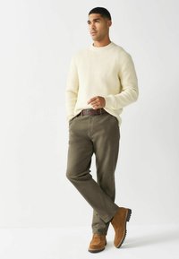 Kreemist kudumist särki, oliivrohelised püksid, pruunist nahast saapad. Mudel poseerib ühe käega taskus, kergelt nõjatudes, minimalistlik ökosysteem.