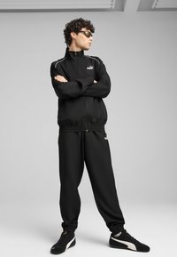 Black tracksuit made of sleek fabric, featuring a zippered jacket with white accents and matching pants, paired with black athletic shoes.