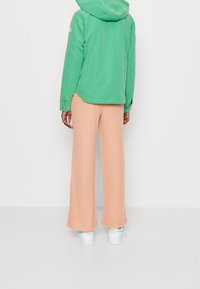 Zielona kurtka z kapturem, brzoskwiniowe szerokie spodnie w prążki, białe sneakersy. Gładki materiał kurtki, teksturowany materiał spodni.