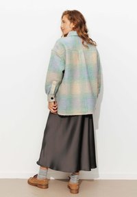 Veste en laine à carreaux de couleurs pastel, avec un col et des poignets boutonnés. Portée sur une jupe en satin foncé, avec des chaussures beiges et des chaussettes grises.