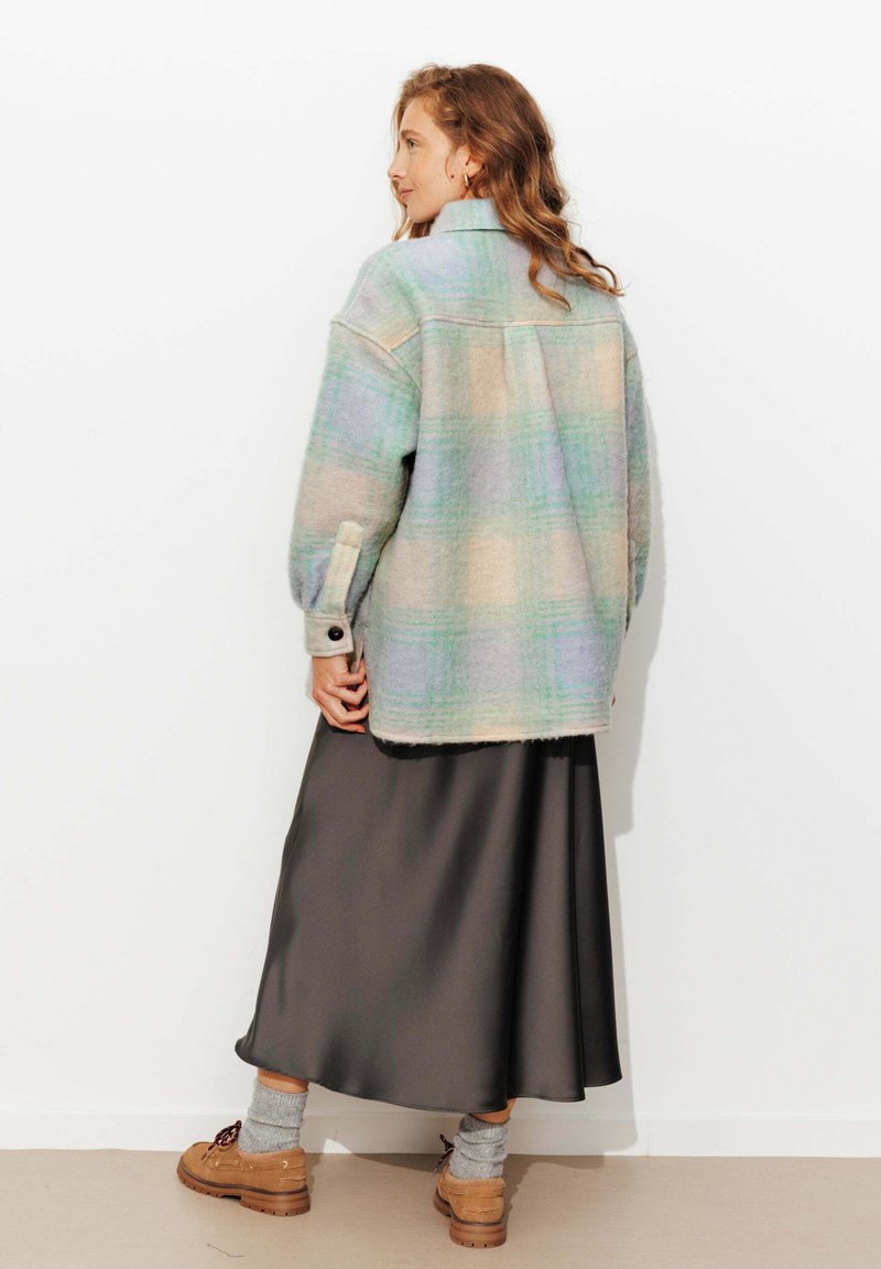 Veste en laine à carreaux de couleurs pastel, avec un col et des poignets boutonnés. Portée sur une jupe en satin foncé, avec des chaussures beiges et des chaussettes grises.