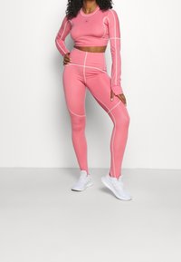 Tajtat, långärmad crop top och högmidjade leggings i solid rosa med vita sömmar. Materialet verkar elastiskt. Bärs med vita skor.