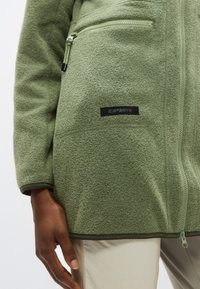 Πράσινο φλις μπουφάν με υφασμάτινη υφή, διαθέτει τσέπη με φερμουάρ και μαύρη ετικέτα μάρκας. Ομαλή λεπτομέρεια φερμουάρ κατά μήκος του μπροστινού μέρους.