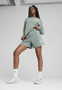 Sweatshirt vert et shorts assortis en tissu doux. Présente le logo PUMA en blanc. Le modèle porte des chaussettes blanches et des baskets de couleur claire.