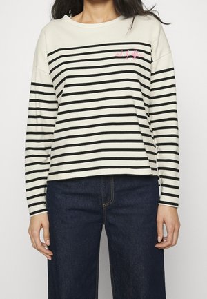 Woman wearing a cream long-sleeve shirt with black horizontal stripes and "out of office" text, paired with dark blue denim jeans.