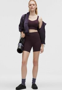 Top corto atlético color burdeos y shorts, combinados con una chaqueta a juego, zapatos negros tipo slip-on y calcetines coloridos. Esterilla de yoga sostenida en la mano izquierda.