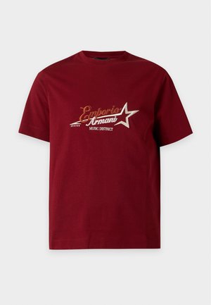 Mand iført beige kasket, rød t-shirt med gyldent stjernemotiv, sorte bukser og sorte sko, stående mod en ensfarvet hvid baggrund.