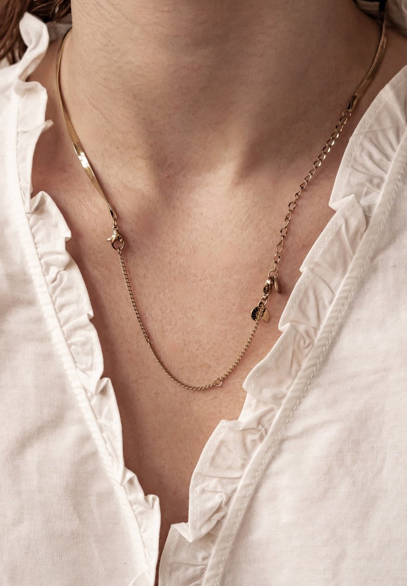 Collier en or avec une chaîne rectangulaire plate et une chaîne plus fine, ornée de petits pendentifs. Porté sur un chemisier blanc à volants.