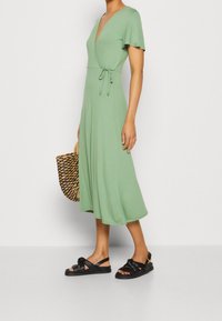 Vihreä kietaisumekko lyhyillä hihoilla, v-pääntiellä ja säädettävällä vyöllä vyötäröllä. Yhdistetty mustiin sandaaleihin ja kudottuun käsilaukkuun.