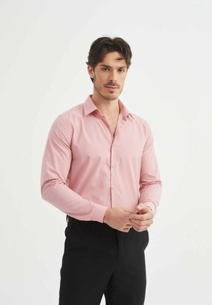 Chemise habillée à manches longues de couleur rose clair avec un col pointu, fermeture à boutons sur le devant et une texture lisse. Associée à un pantalon noir.