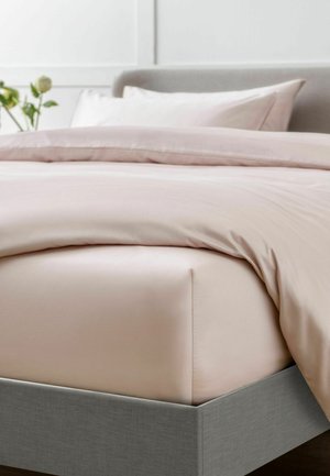 Bett mit hellbeigem Spannbettlaken, passender Bettdecke, Kissen, grau gepolstertem Rahmen und einer weißen Blume im Hintergrund an einer hellen Wand.