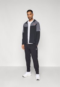 Αθλητική εμφάνιση με μαύρο και γκρι τζάκετ με φερμουάρ, λευκό μπλουζάκι, μαύρο παντελόνι και λευκά αθλητικά παπούτσια. Περιλαμβάνει λογότυπα και ομαλή υφή.