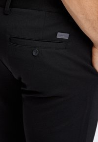 Svarta byxor med texturerat tyg, med en bakficka och en svart knapp. En liten grå etikett syns på fickan.