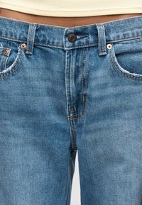 Κοντινό πλάνο της μέσης που φορά μπλε τζιν με φθαρμένες μπροστινές τσέπες και μπεζ ριμπ τοπ που έχει μπαίνει μέσα.