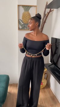 Zwarte off-shoulder top gecombineerd met losse zwarte broek, houdend een gouden charmeketting met verschillende kleurrijke hangers.