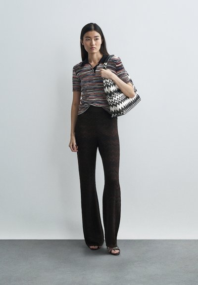 Woman wearing patterned short-sleeve top, black sheer zigzag pants, holding black-and-white zigzag tote bag, standing on gray floor.