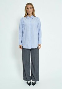 Camicia button-up a righe blu con maniche lunghe, abbinata a pantaloni larghi grigi e scarpe nere con punta affilata.