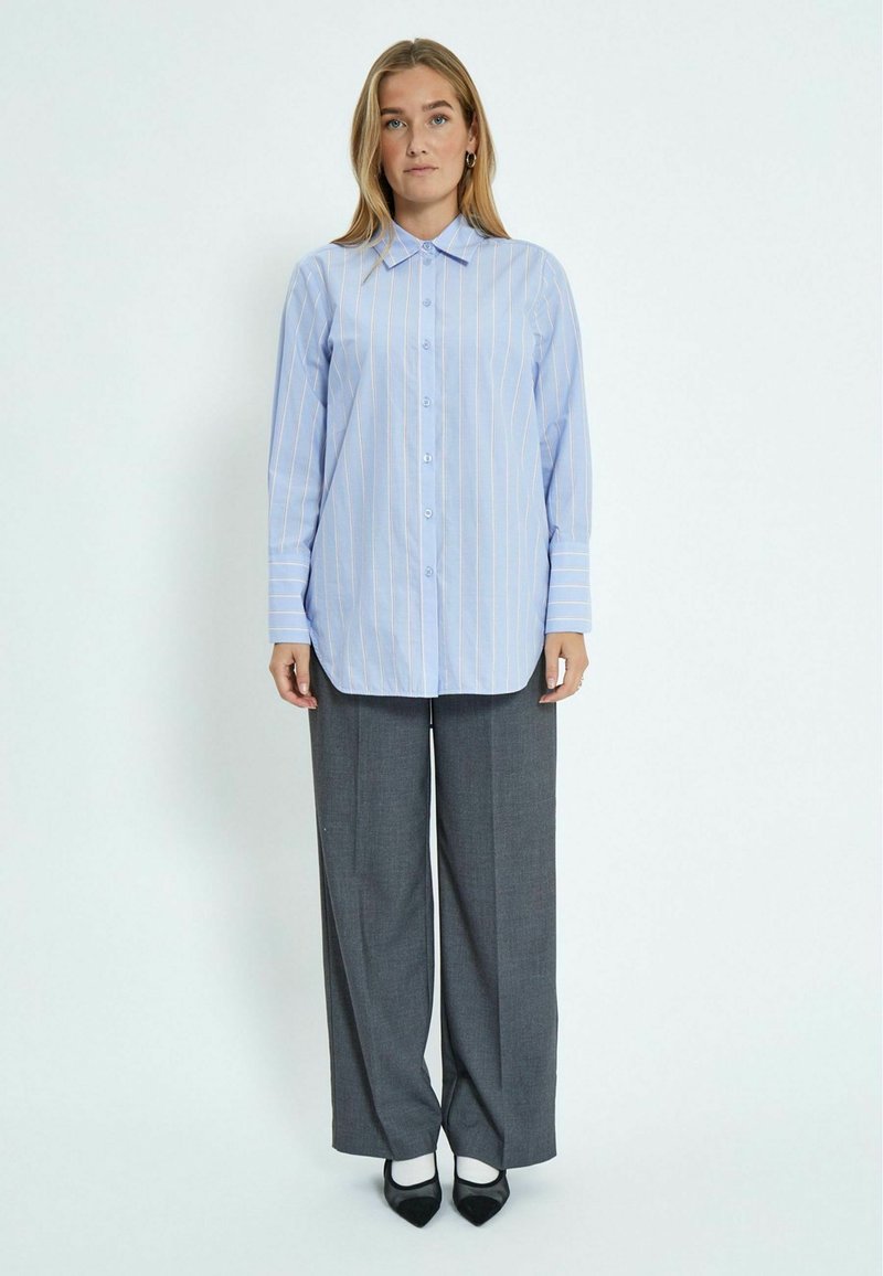 Camicia button-up a righe blu con maniche lunghe, abbinata a pantaloni larghi grigi e scarpe nere con punta affilata.