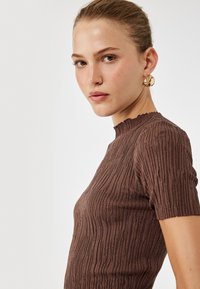 Brun ribbet kortærmet top med høj hals og tekstureret stofdetalje. Model med hoops øreringe, vinkelret udsigt.
